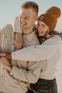 Happy couple enjoying a snowy day in the golden sunshine, the girl has her arms wrapped around the guys shoulders, they are smiling and laughing