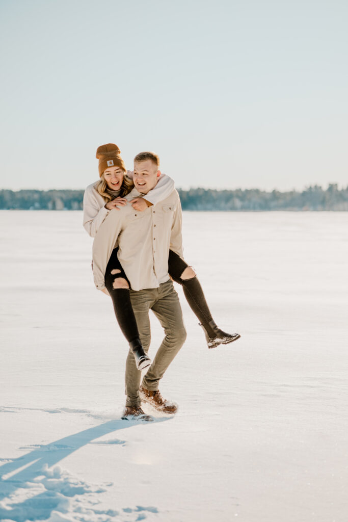 Happy couple enjoying a snowy day, with the guy giving his fiancée a piggyback ride, both smiling and laughing
