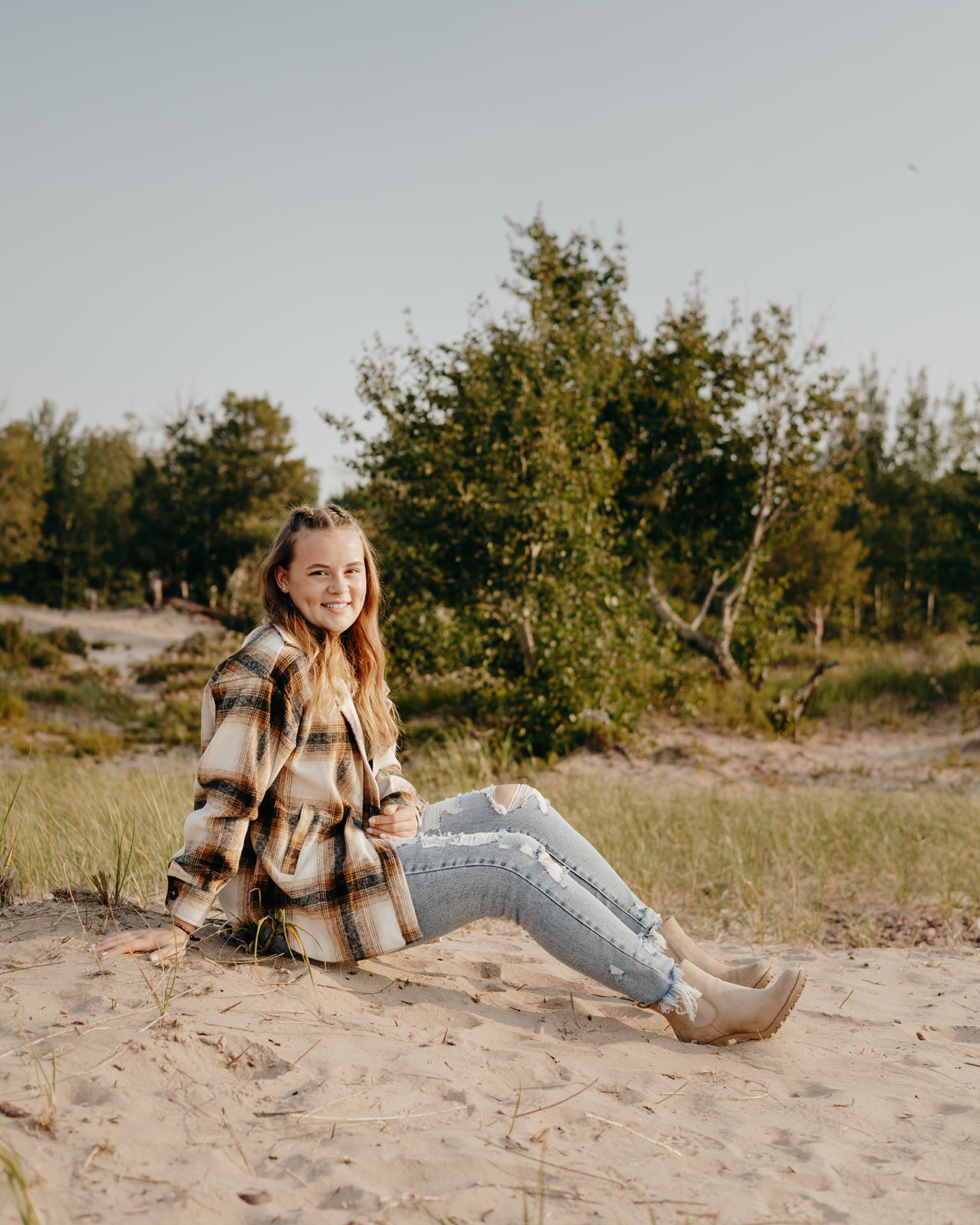 Abi leans on a sand dune in a cozy flannel shirt during her senior photos in Eagle River, Michigan