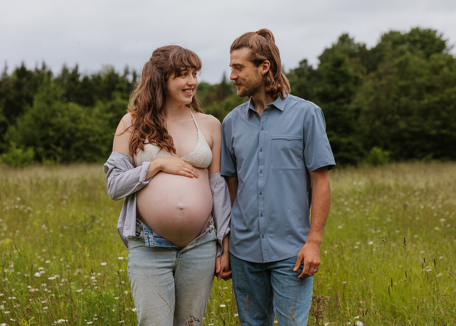 Husband and wife share a smile in an open grassy field during their summer maternity session in Hancock Michigan