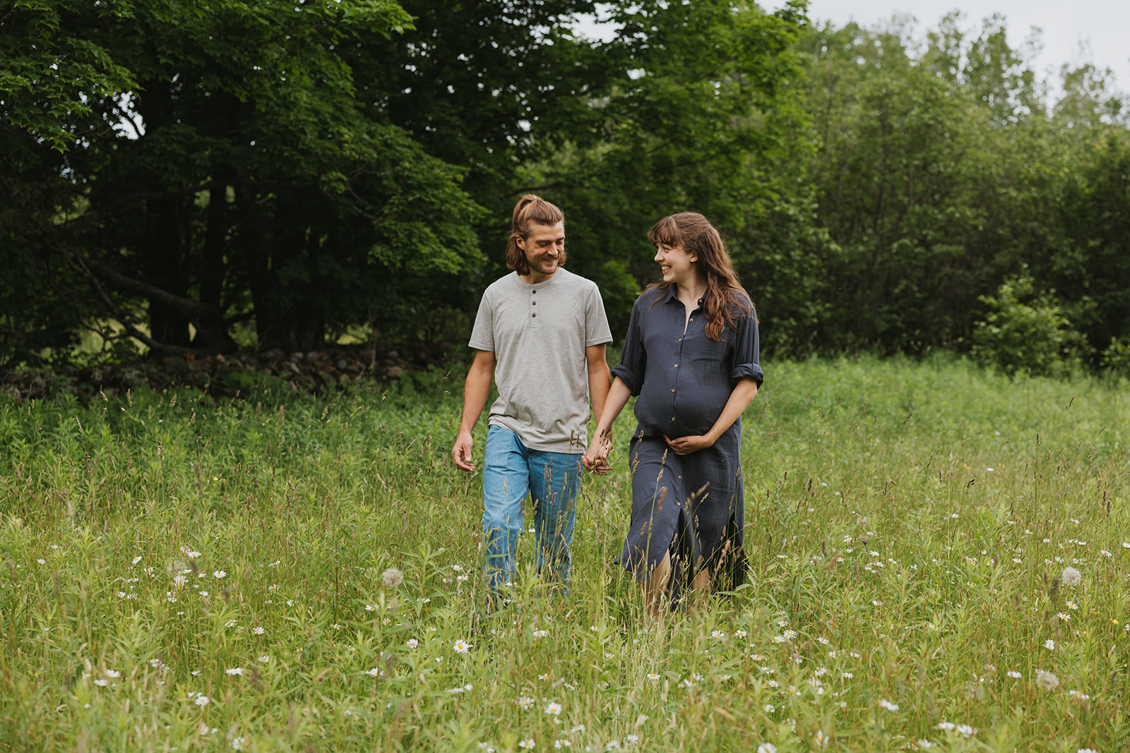 Couple walk together though a grassy field during their summer maternity session in Hancock, Michigan